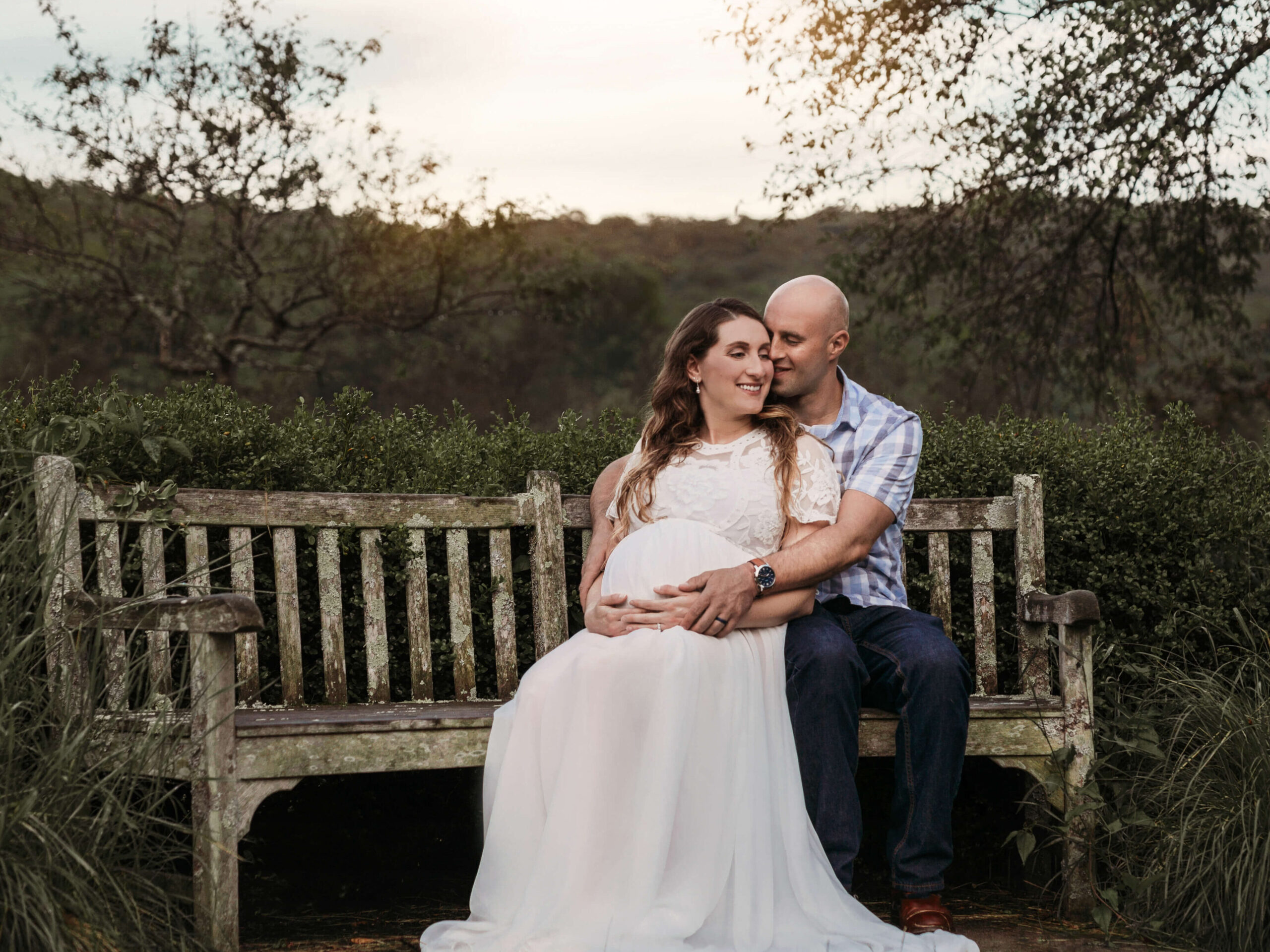 northern virginia pregnancy and maternity photographer capturing a pregnant woman wearing a white maternity gown and her husband sitting on a bench at sunset in a park in Loudoun County VA