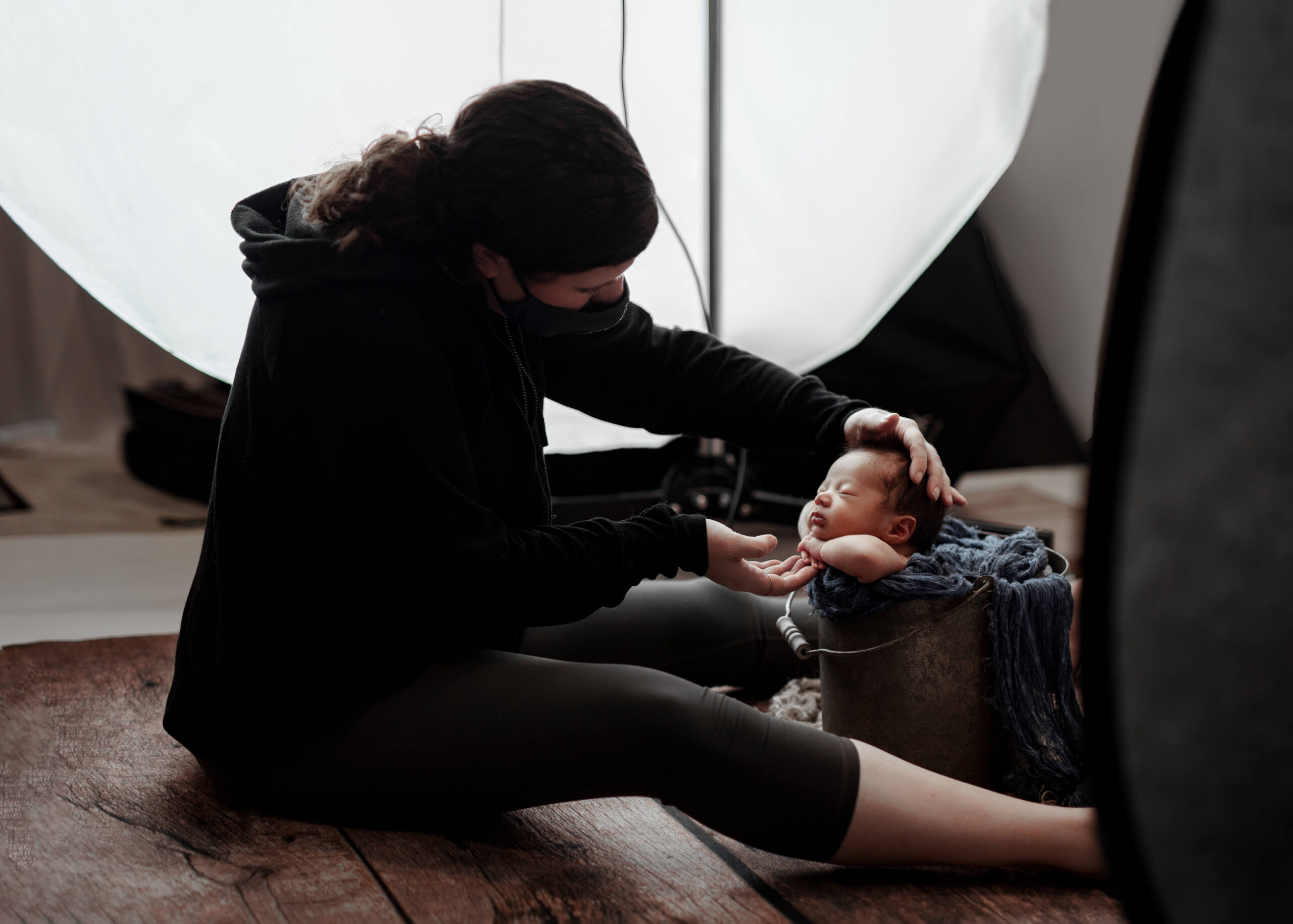 Newborn Baby Photographer wearing a mask for COVID protocols for Newborn Photography while soothing and posing a newborn baby posed in a bucket