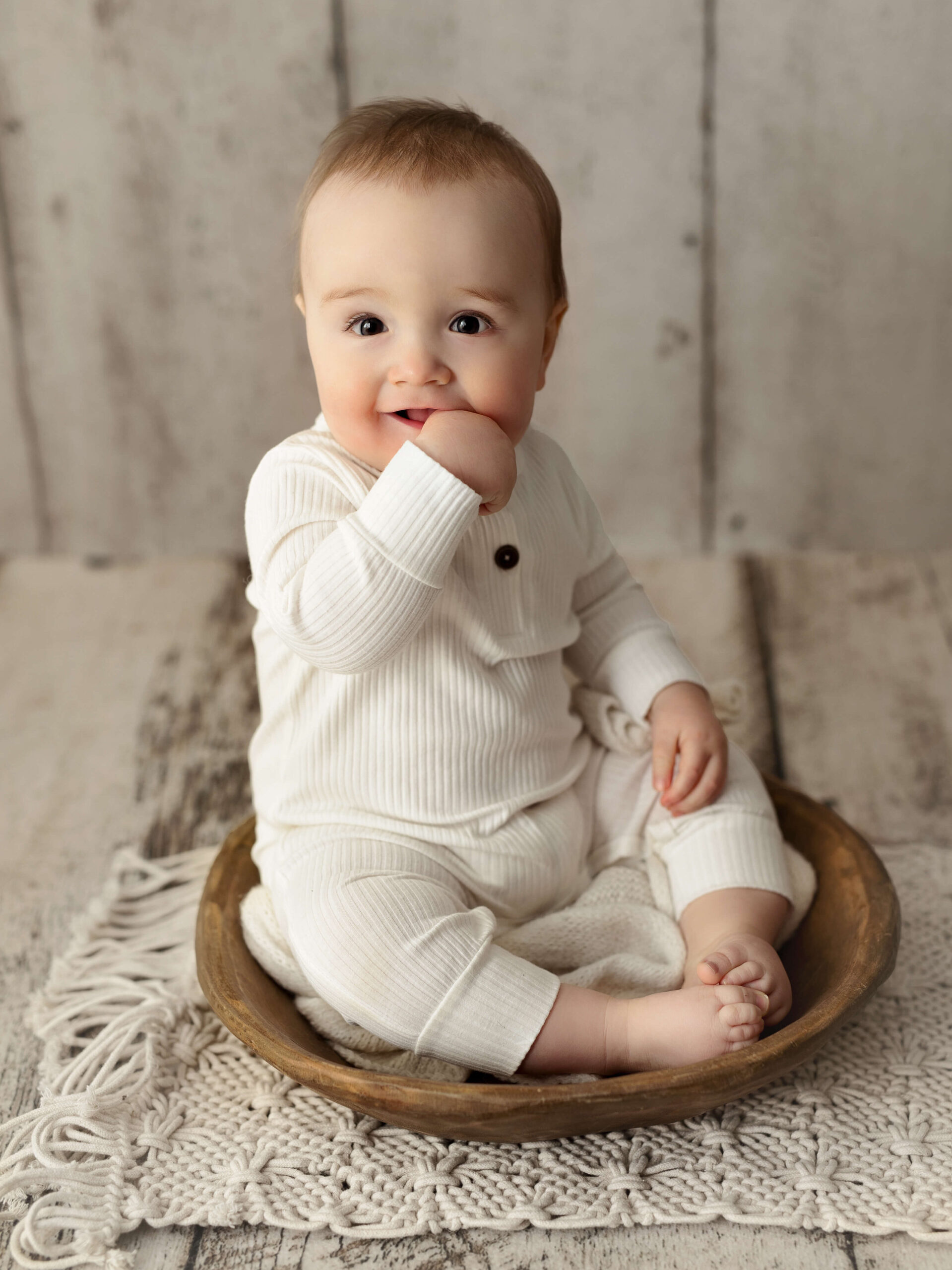 sitter photo taken by a Loudoun VA baby photographer, 6 month old in a white onesie sitting in a wooden bowl with his thumb in his mouth at a milestone baby photo shoot