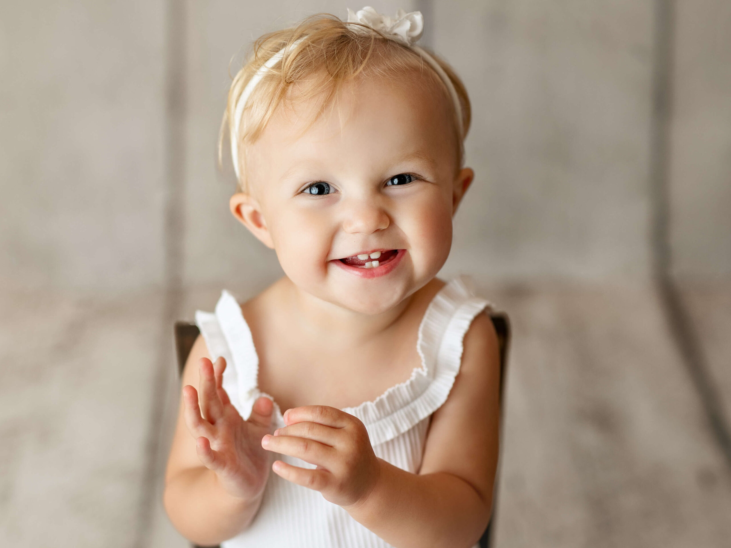 milestone photo taken by a Loudoun VA baby photographer, 9 month old in a white outfit sitting in a wooden chair clapping her hands at a milestone baby photo shoot