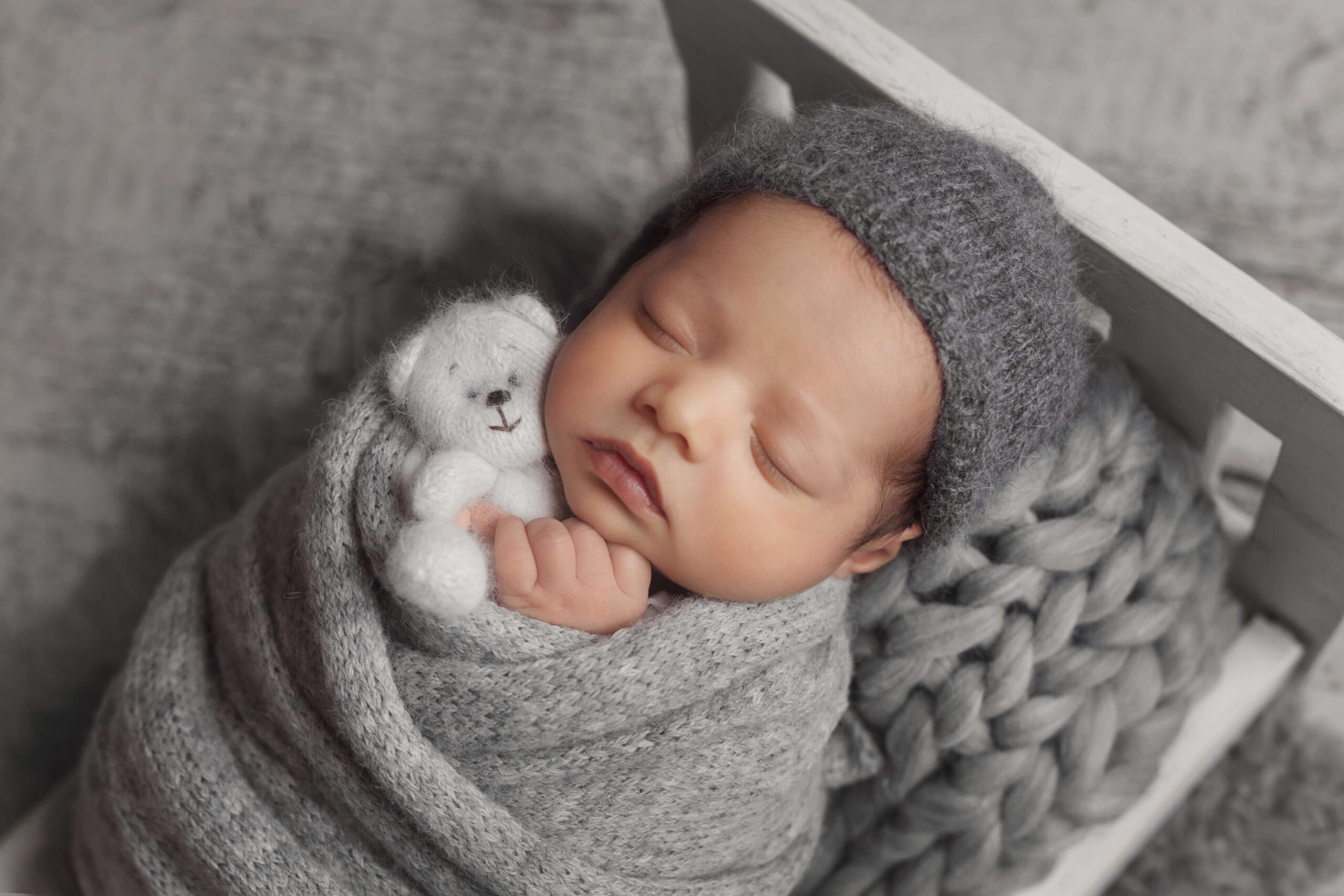 newborn baby sleeping while swaddled in a grey knit wrap wearing grey sleepy cap holding a white knit bear while laying in a white wooden bed, soothing babies during photo session, Serling VA newborn photographer