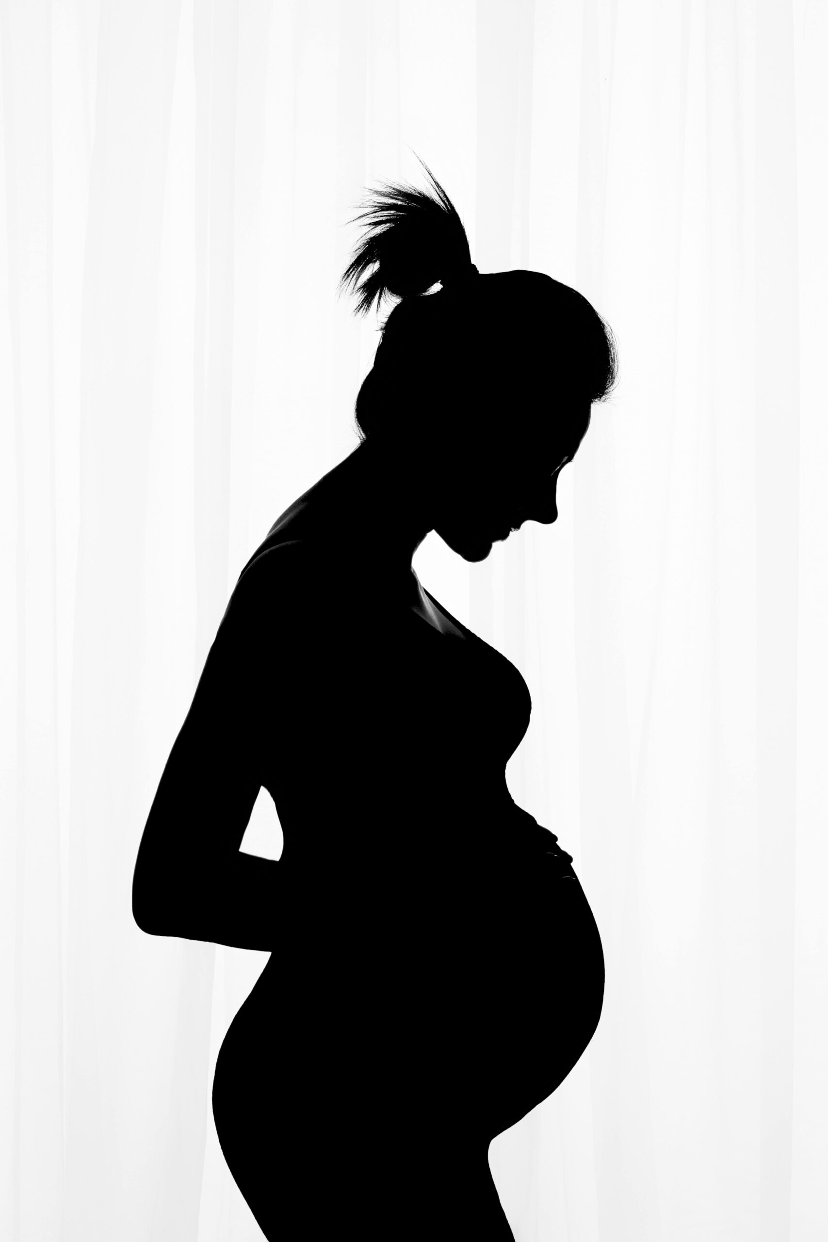 black and white photo of a pregnant woman standing in front of a white back-lit curtain taken by a maternity photographer, When to Take Maternity Photos