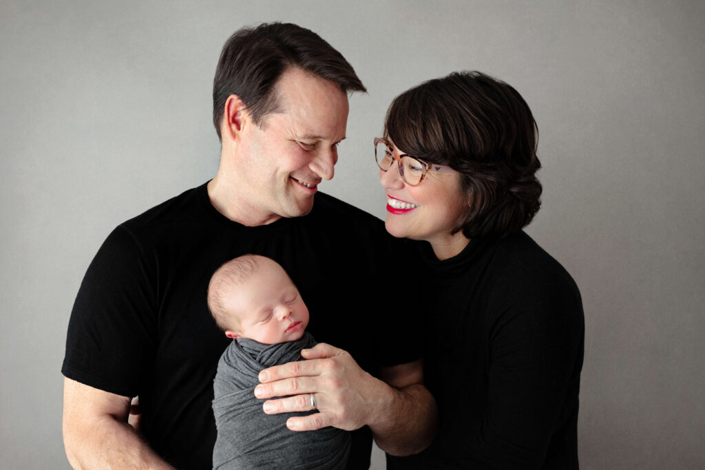 mom and dad smiling at each other wearing black outfits while dad is holding their newborn son in a grey backdrop for their family newborn pictures