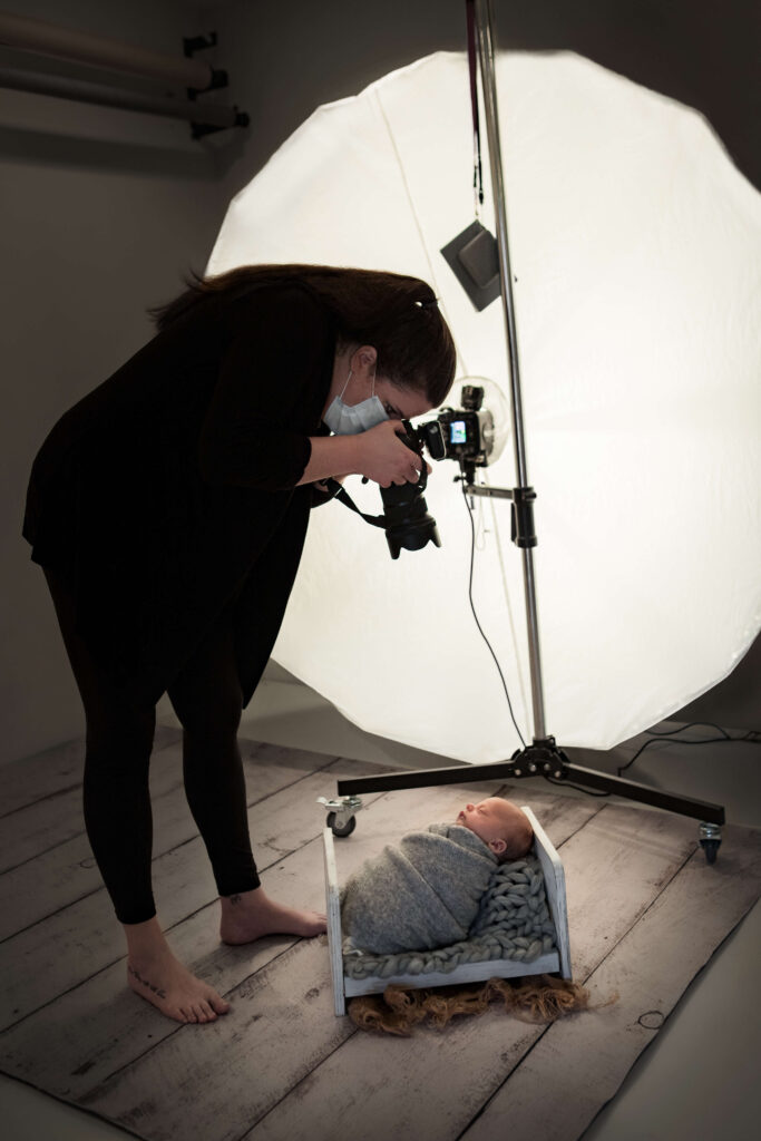 photographer wearing a mask for COVID protocols for Newborn Photography while taking a photo of a baby swaddled in a wooden bed
