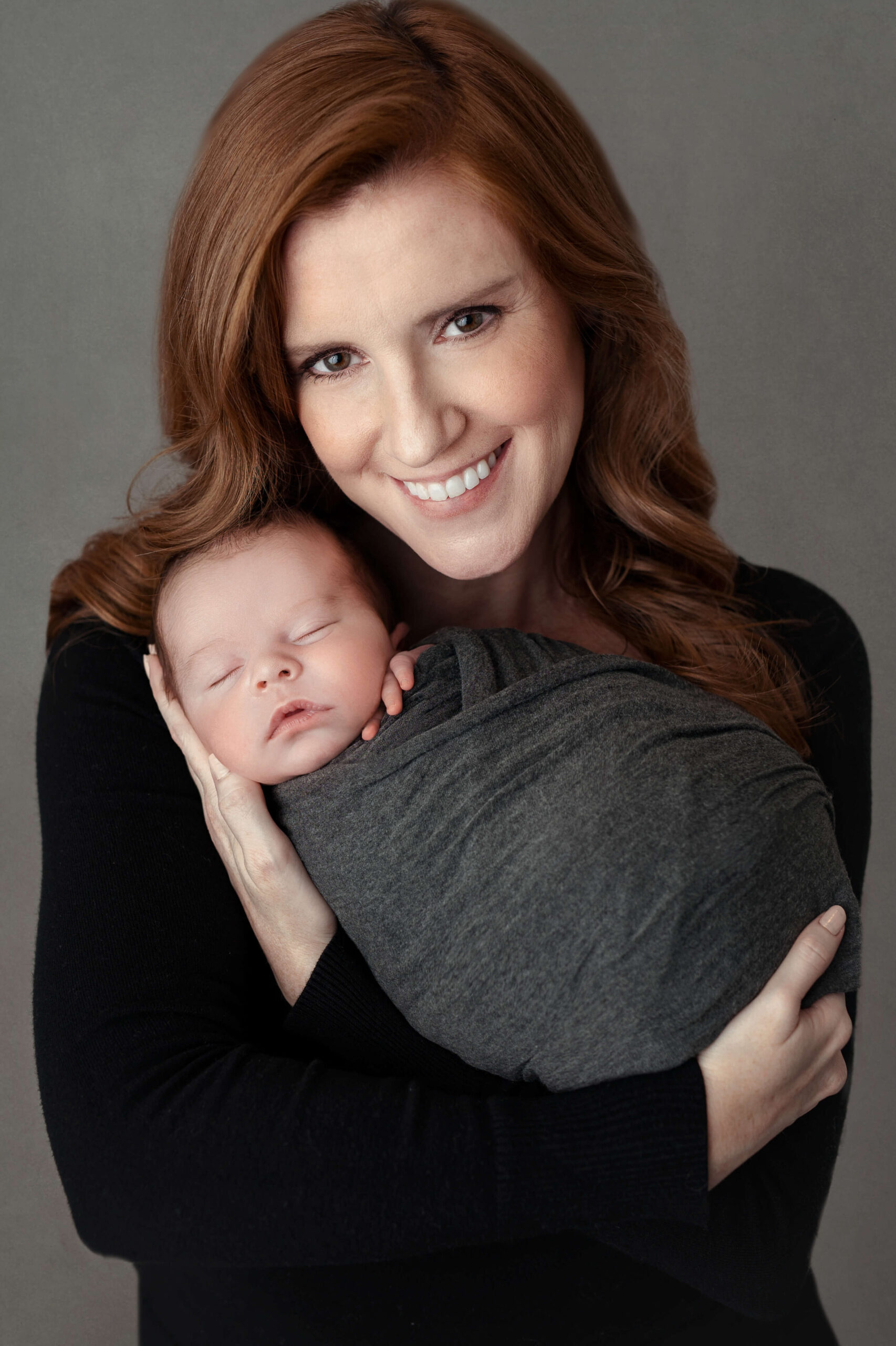 new mother in a simple comfortable outfit smiling holding her newborn boy who is swaddled in grey on a grey backdrop for her parent pictures at her newborn session