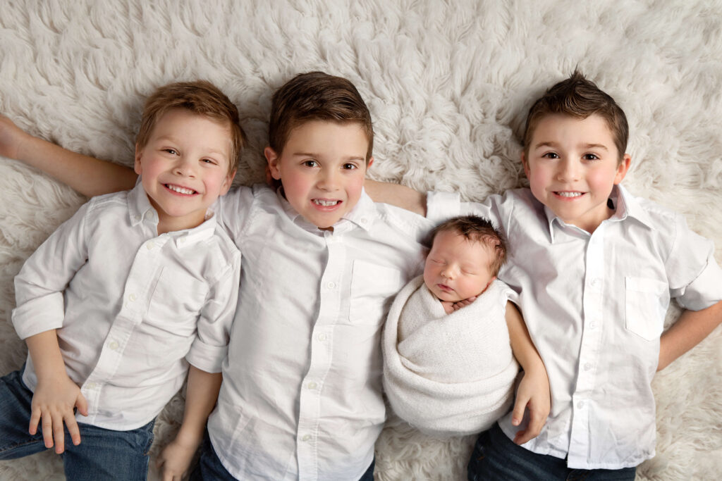 three big brothers wearing white button-up shirts with their newborn brother laying together on a white fur rug for their family newborn pictures in a newborn session