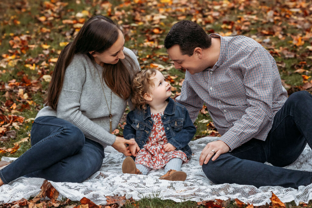 Fall Family photo shoot in a park in Leesburg VA, mom and dad sitting on a blanket in the all leaves with their toddler sitting in between them.
