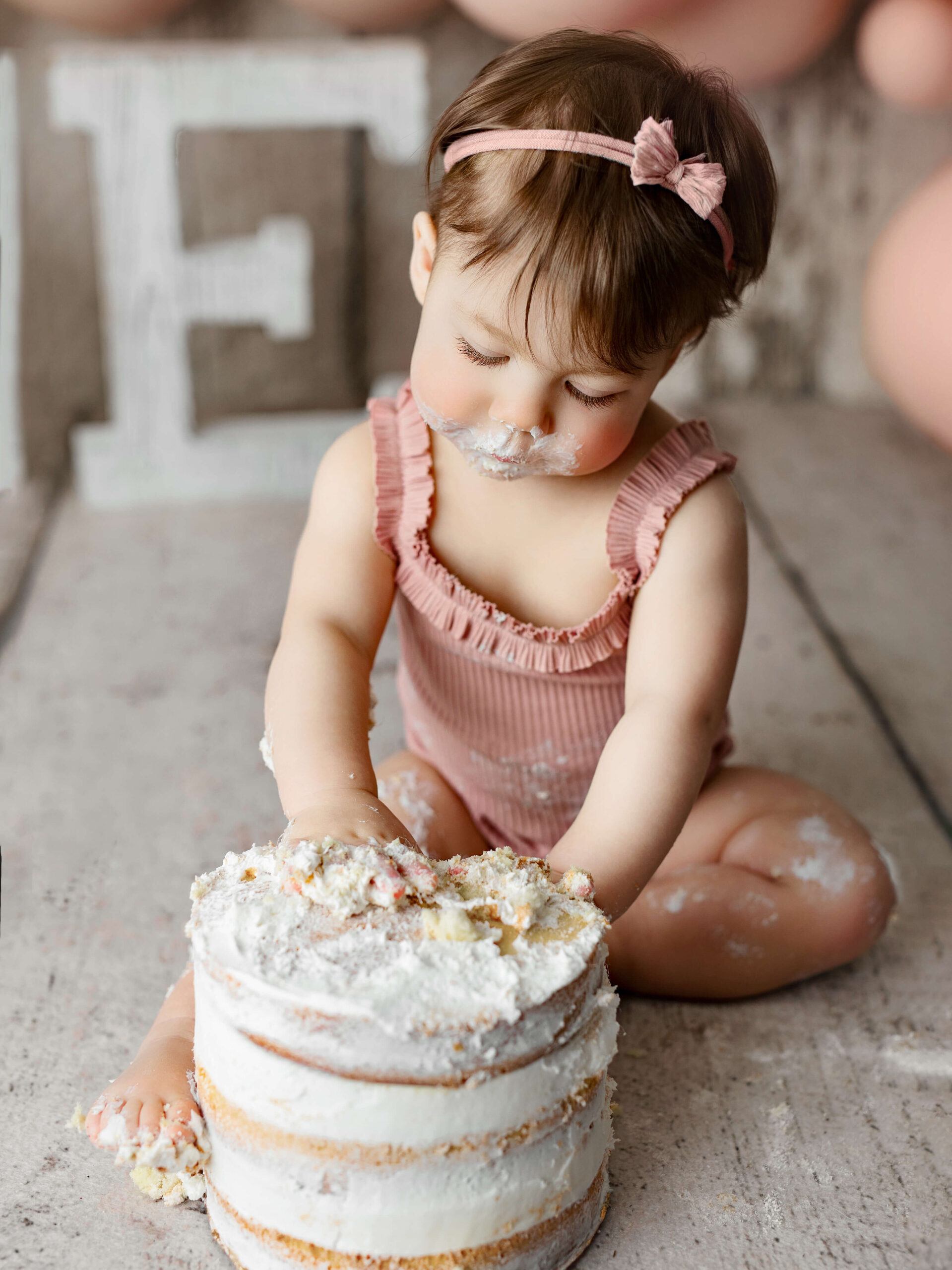 cake smash photo taken by a Loudoun VA baby photographer, one year old in a pink outfit sitting playing with her birthday cake at a milestone or cake smash baby photo shoot