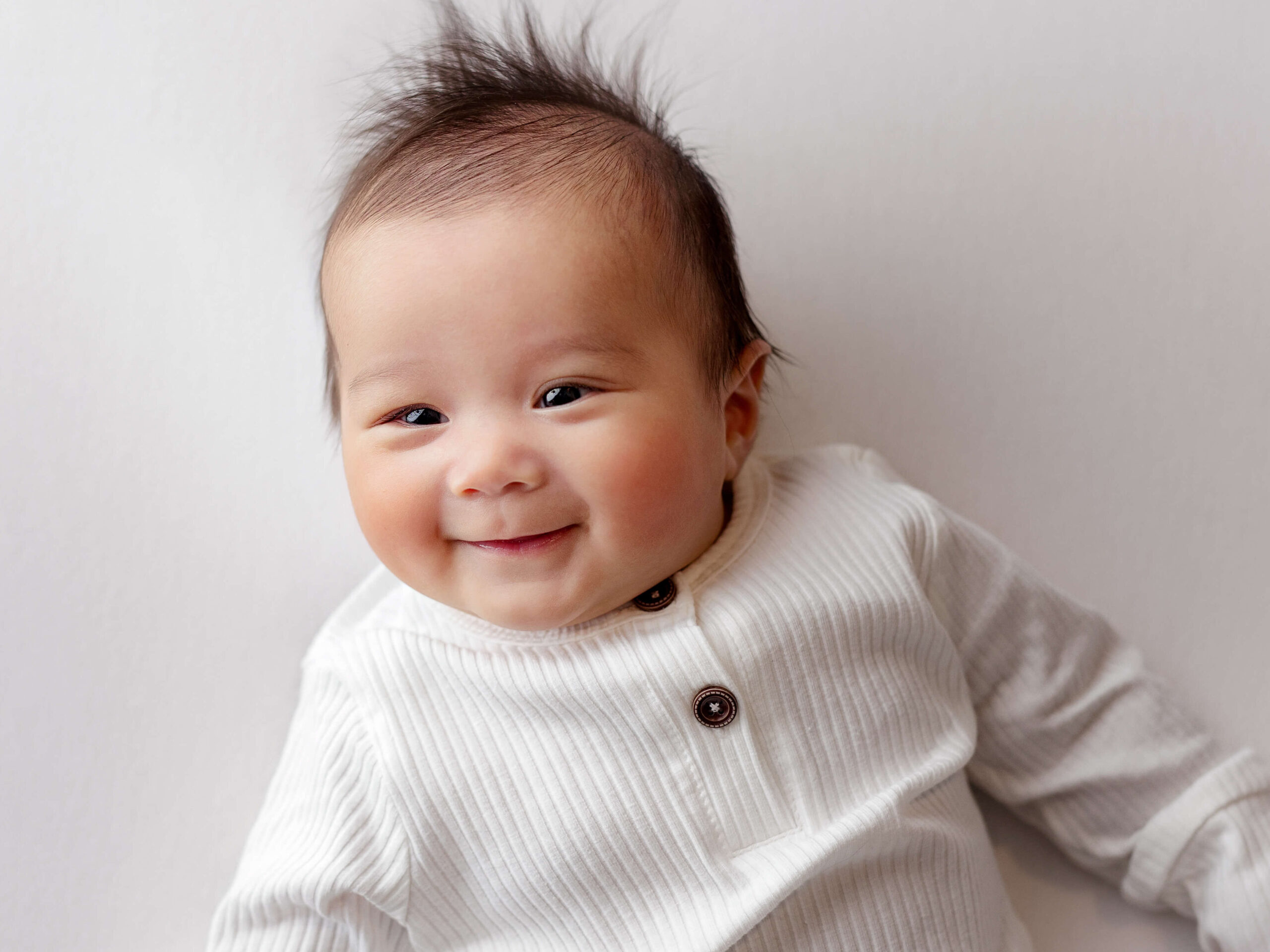 3 month old photo taken by a Loudoun VA baby photographer, three month old baby in a white onesie laying on a white backdrop smiling at a baby photo shoot