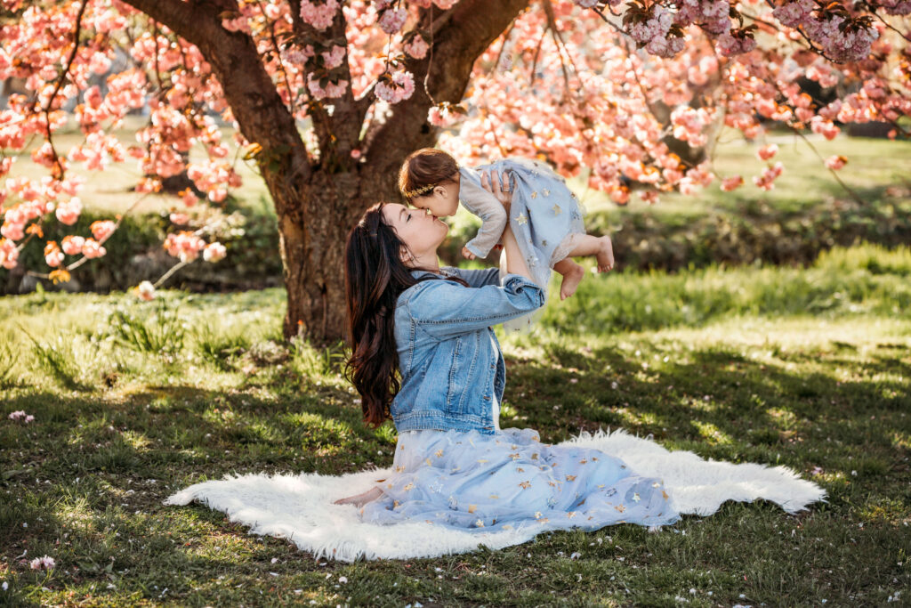 Mom holding her daughter up in the air kissing her in front of a tree in the spring at an outdoor family photo shoot at golden hour.