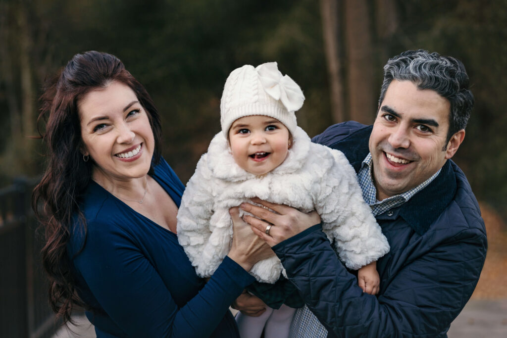 Winter family photography session in Great Falls VA, happy mom and dad wearing navy blue holding their daughter who is laughing wearing a white hat and jacket. wearing