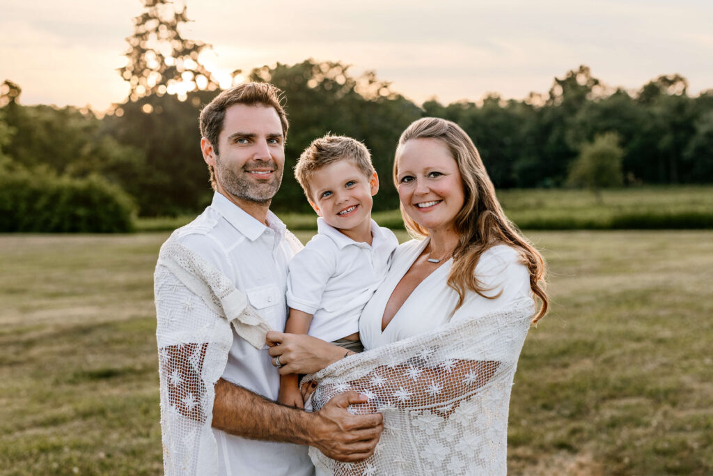 Family photo shoot taken at sunset in the summer in a park in leesburg VA, mom and da holding their son in between them with a blanket around the family wearing white