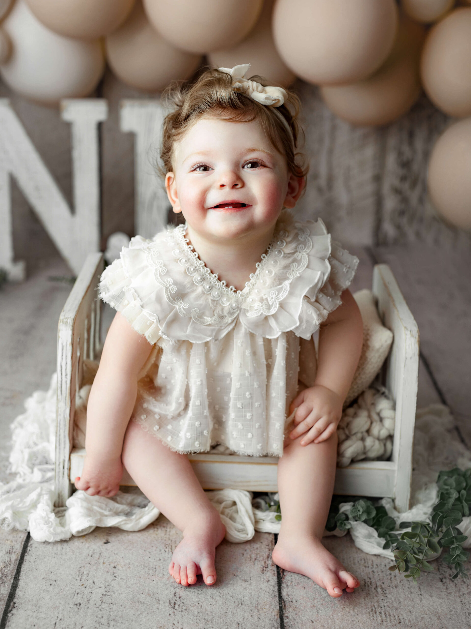 toddler in white outfit in a wooden bed smiling at a Cake Smash Photo Shoot at a Fairfax VA Toddler Photography Studio