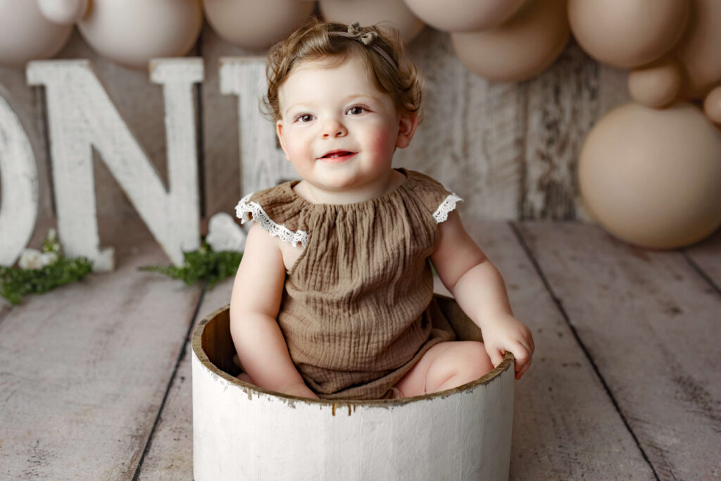 toddler in brown onesie in a white wooden bucket at a Cake Smash Photo Shoot at a Fairfax VA Toddler Photography Studio