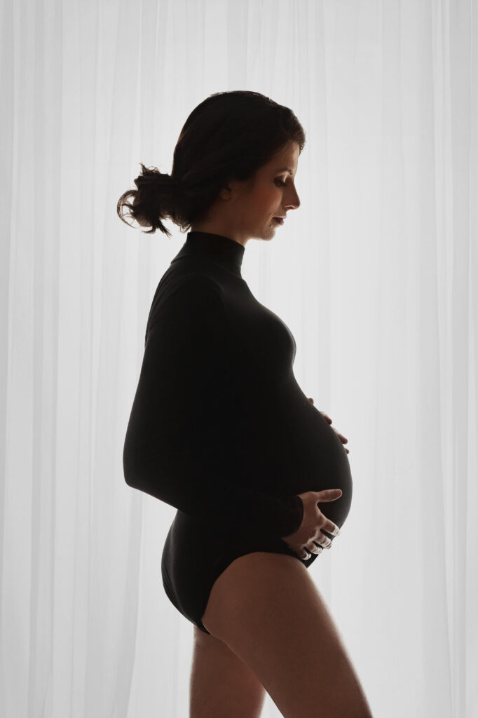 pregnant woman wearing a black body suit standing in front of white sheer curtains at her maternity session in northern va