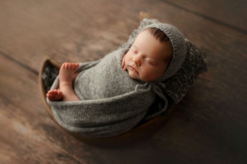 newborn in a grey knit wrap and bonnet in a moon wooden bowl taken by a Northern Virginia Newborn Photographer