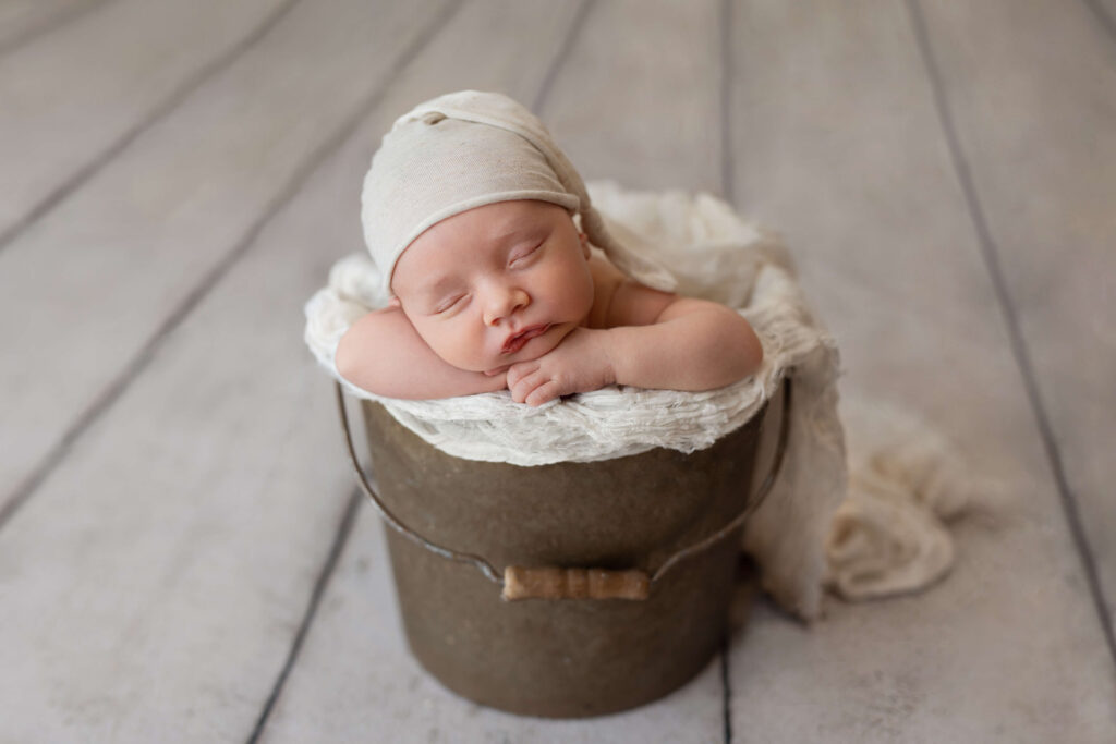 newborn baby in a bucket with a cream sleepy cap taken by a ashburn va newborn photographer