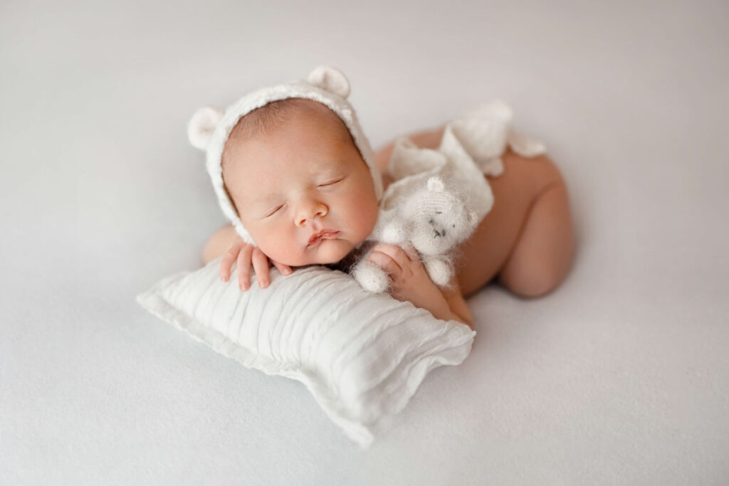 newborn baby naken on a white background with a bear bonnet and bear stuffie on white pillow taken by an ashburn va newborn photographer