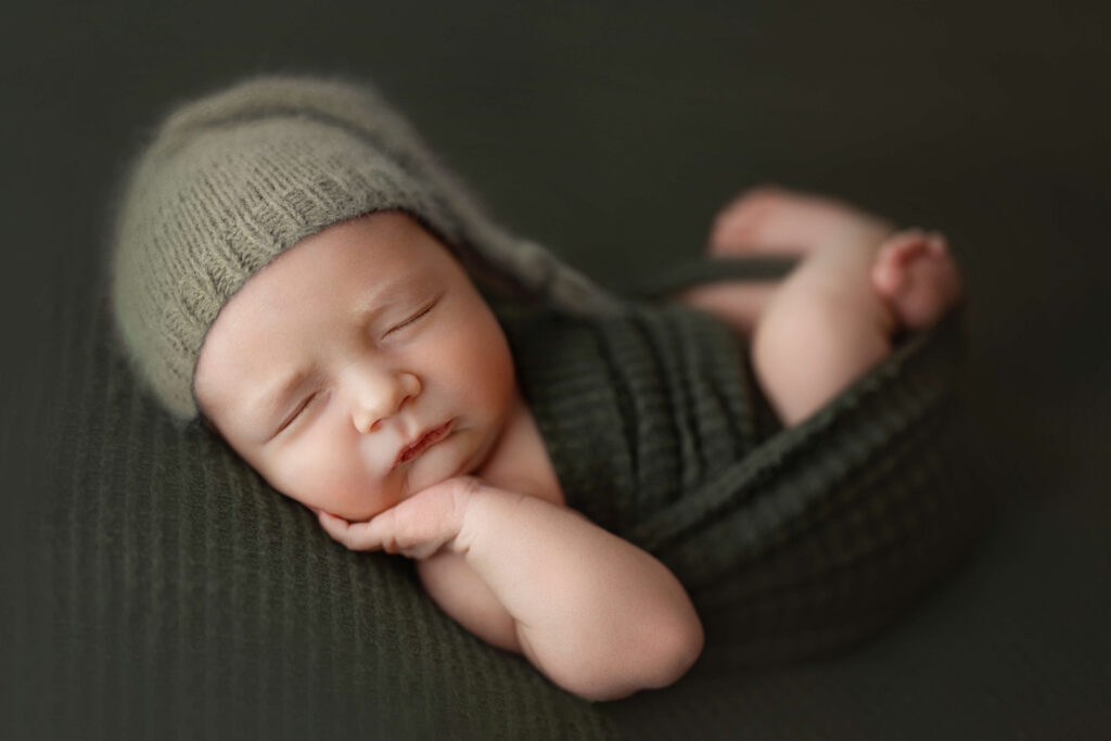 newborn baby with hand on his cheek on a green backdrop with green sleepy cap