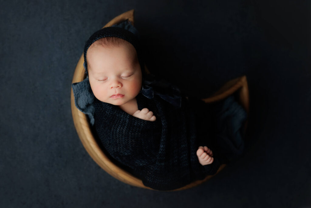 newborn  in a moon bowl with a blue wrap and bonnet on a navy blue backdrop