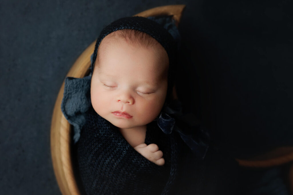 newborn  in a moon bowl with a blue wrap and bonnet on a navy blue backdrop